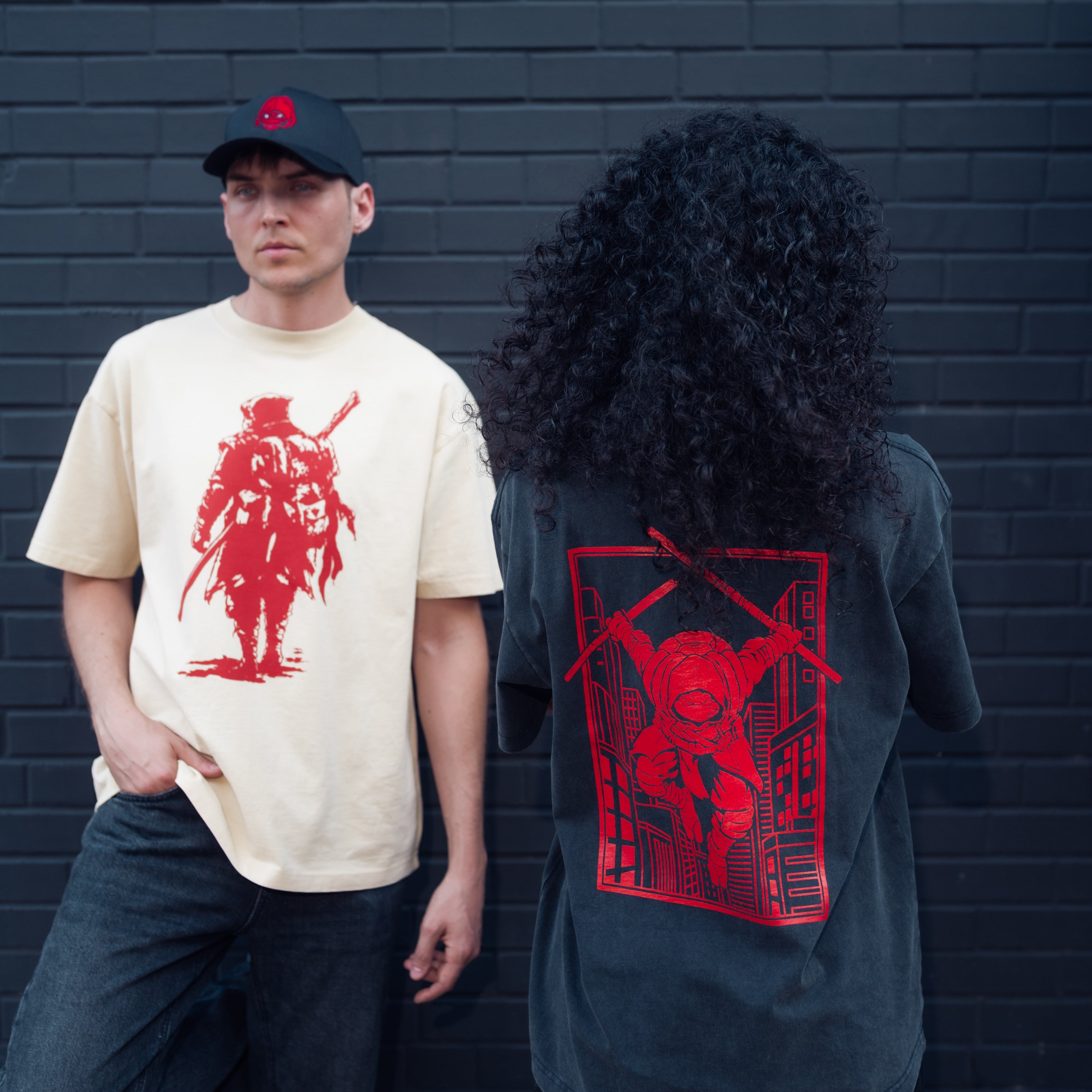 Two people wearing t-shirts with red teenage mutant ninja turtles graphic designs against a dark brick wall.
