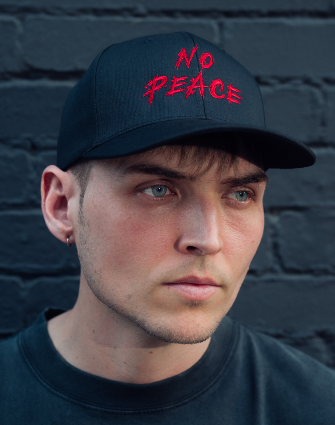 Person wearing a black cap with 'No Peace' in red text against a dark brick wall.